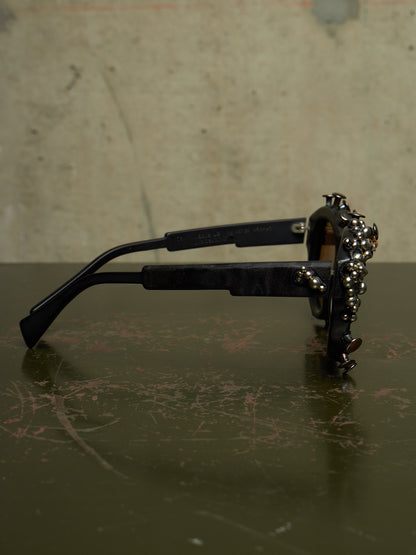 Black sunglasses with an ornate, beaded gold metal frame, resting on a dark scratched tabletop against a textured beige wall.