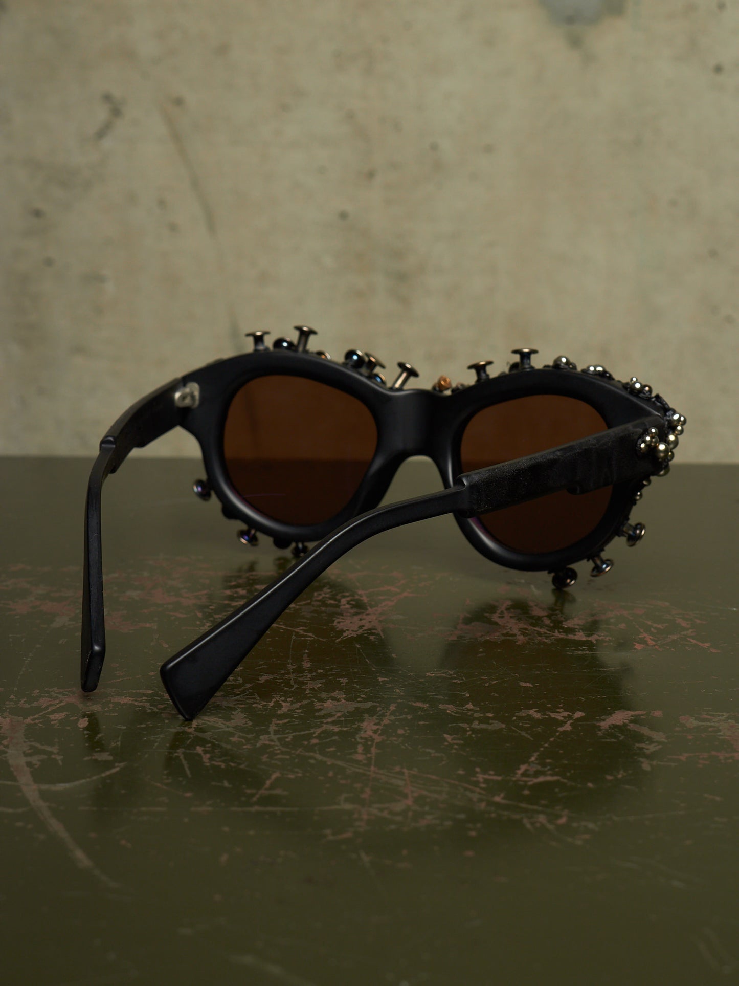 Black sunglasses with brown lenses and decorative metal studs and screws around the frame, resting on a dark textured surface against a beige background.