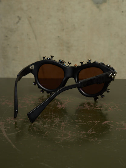 Black sunglasses with brown lenses and decorative metal studs and screws around the frame, resting on a dark textured surface against a beige background.