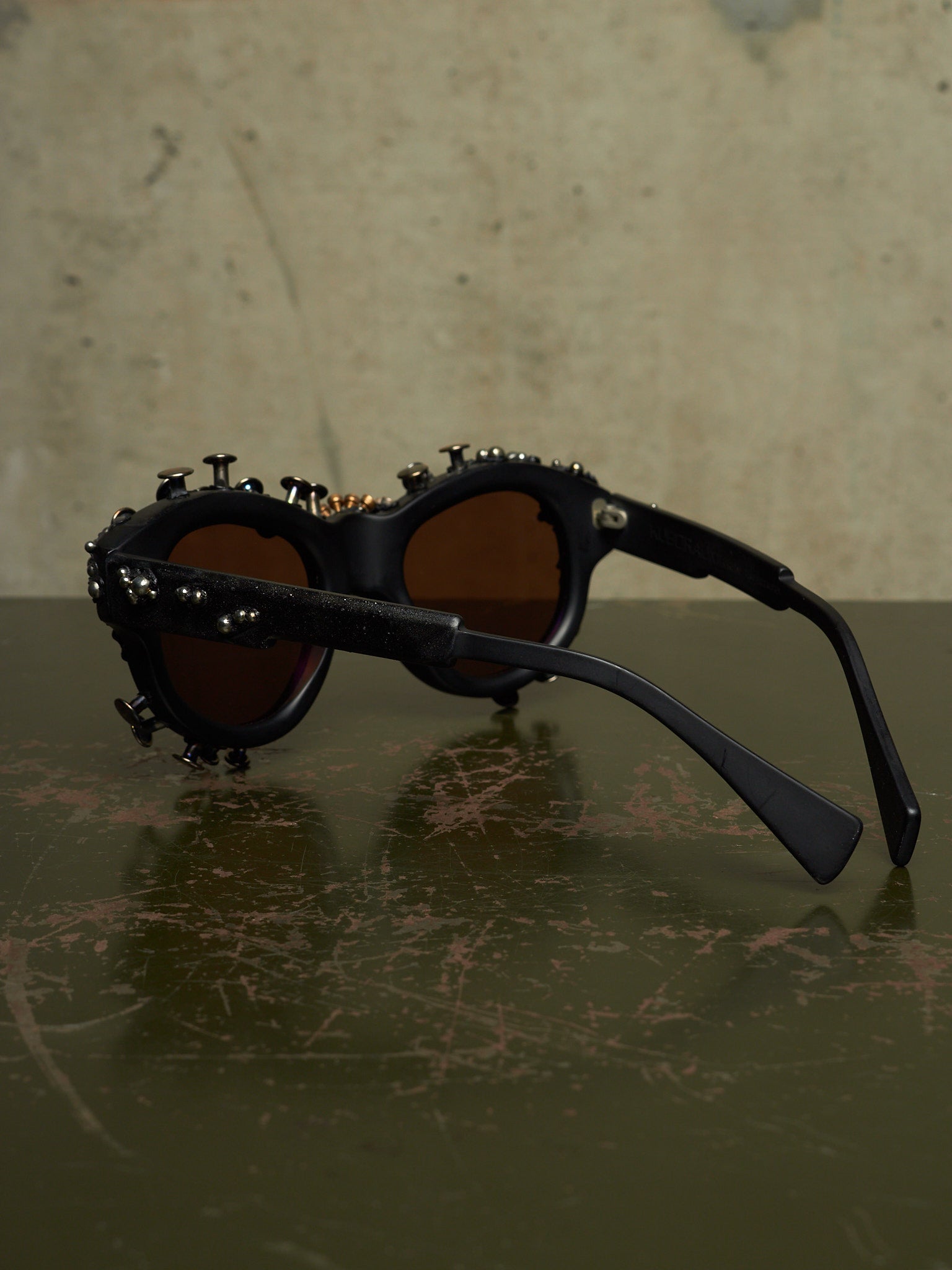 Black studded sunglasses with dark brown lenses, resting on a dark reflective surface against a textured beige background.
