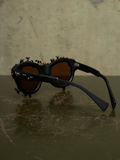 Black studded sunglasses with dark brown lenses, resting on a dark reflective surface against a textured beige background.