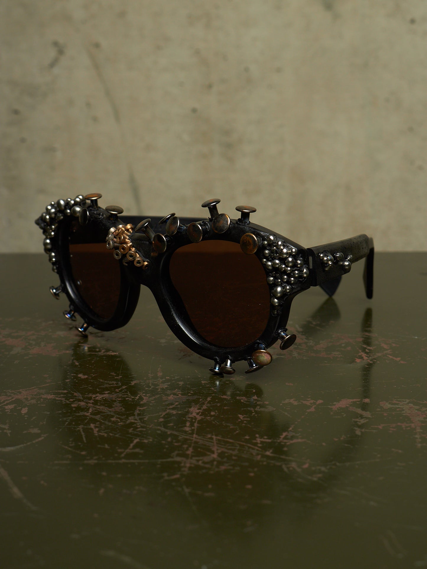 Decorative black sunglasses with dark brown lenses, adorned with metallic studs, rivets, and small gold-toned embellishments, resting on a dark reflective surface against a textured beige background.