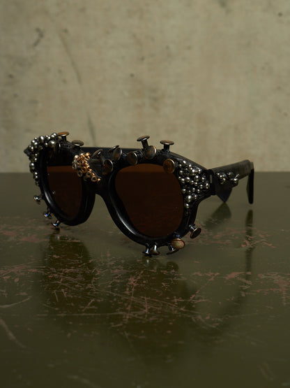 Decorative black sunglasses with dark brown lenses, adorned with metallic studs, rivets, and small gold-toned embellishments, resting on a dark reflective surface against a textured beige background.