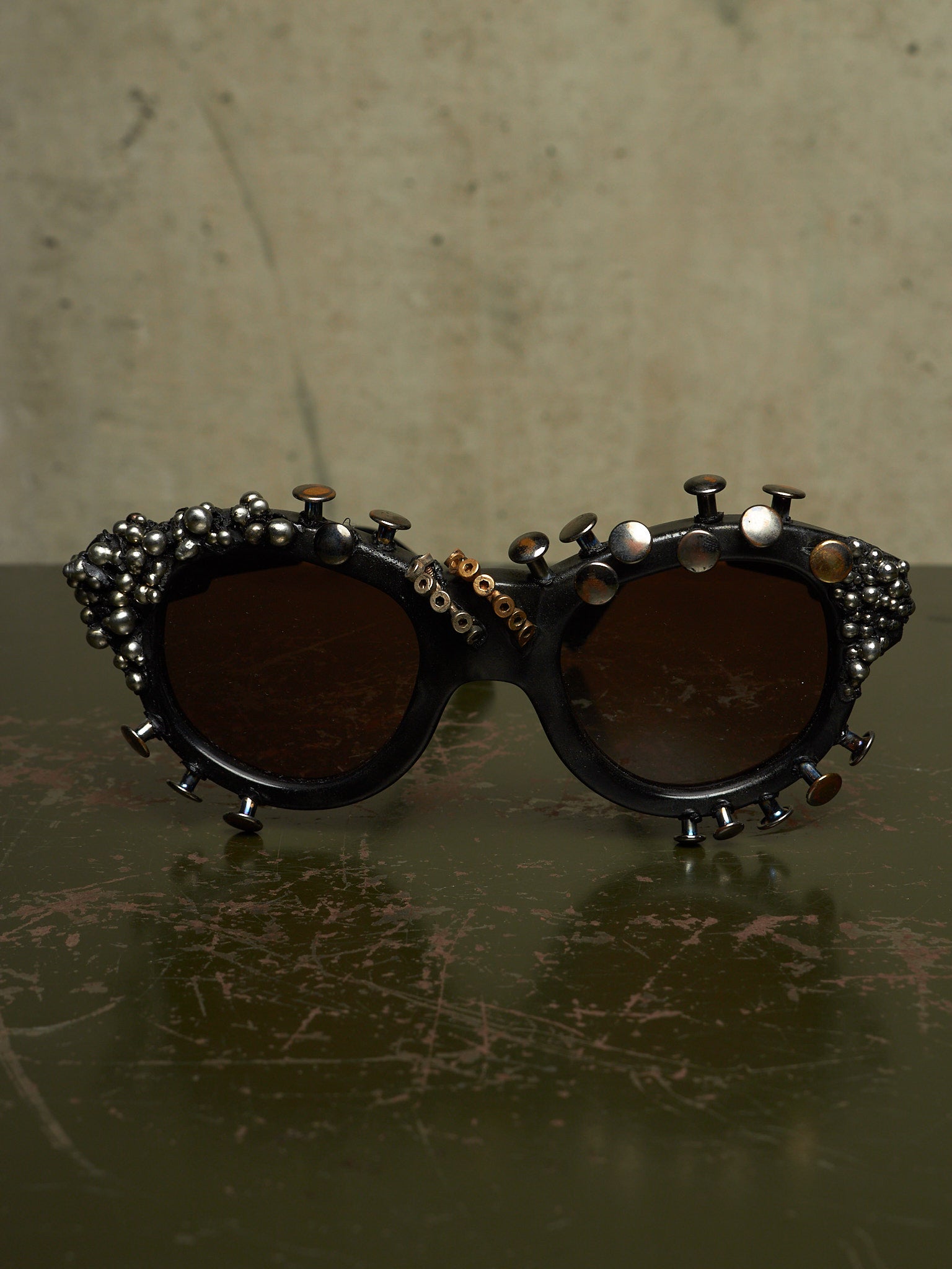 Stylized round sunglasses with dark brown lenses and an ornate black frame decorated with metallic studs, beads, and small metal spikes, displayed on a dark reflective surface against a textured beige background.
