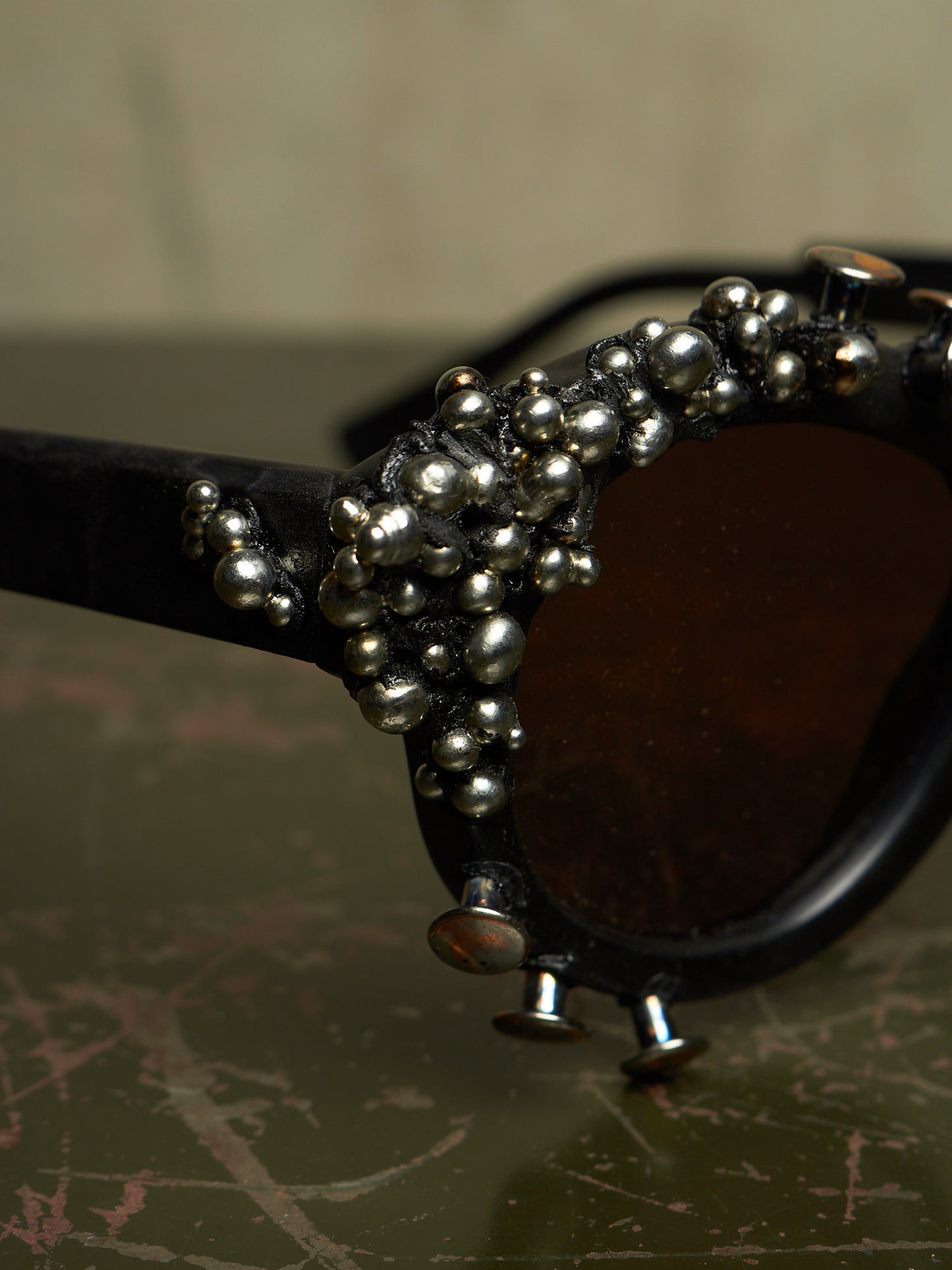 Close-up of a pair of black sunglasses with dark brown lenses and an ornate cluster of silver metallic beads and small studs along the frame, resting on a textured dark green surface.