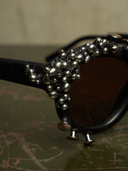 Close-up of a pair of black sunglasses with dark brown lenses and an ornate cluster of silver metallic beads and small studs along the frame, resting on a textured dark green surface.