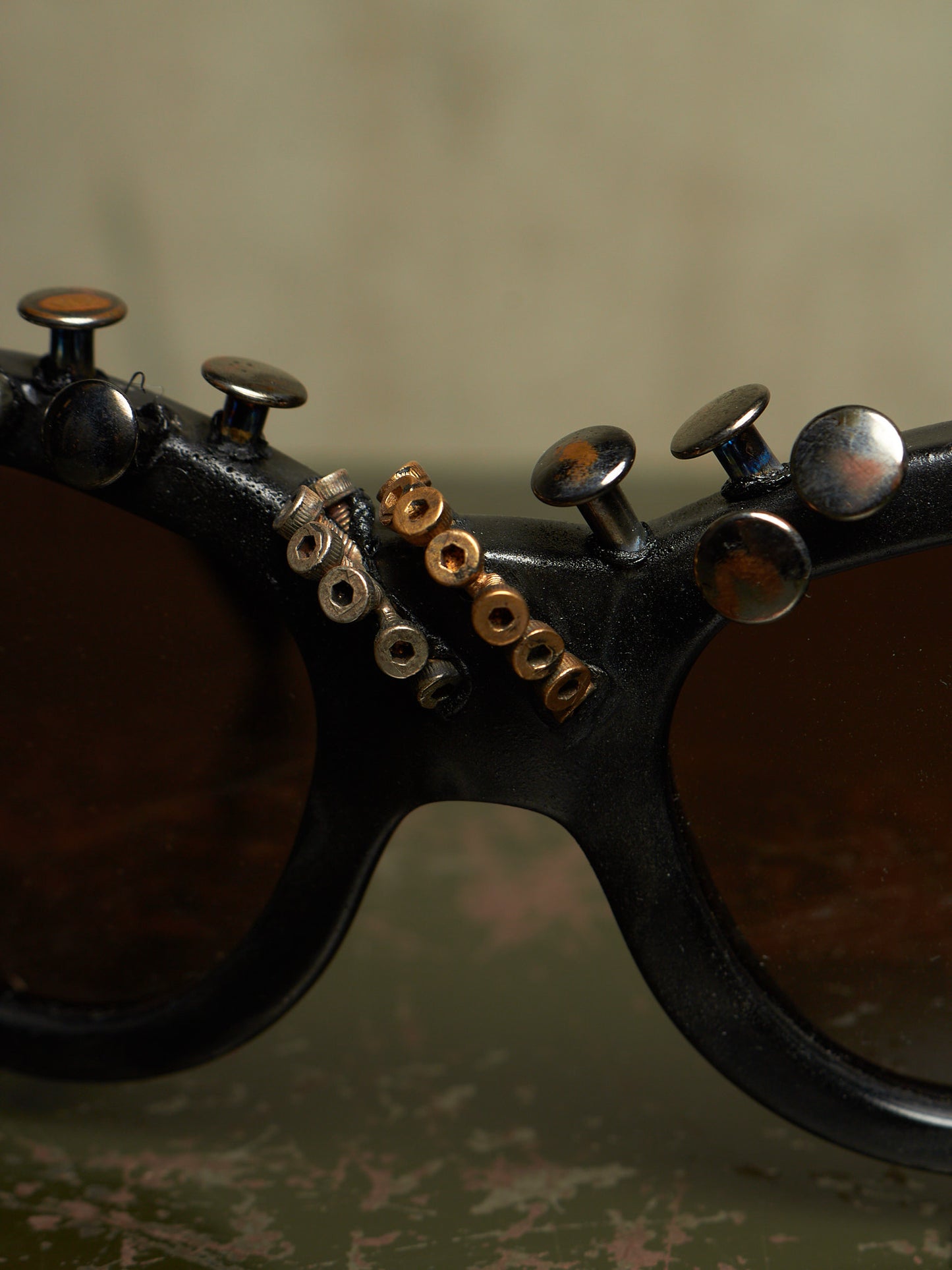 Close-up of a pair of dark sunglasses with decorative metallic rivets and screws along the top frame, set against a textured beige background.
