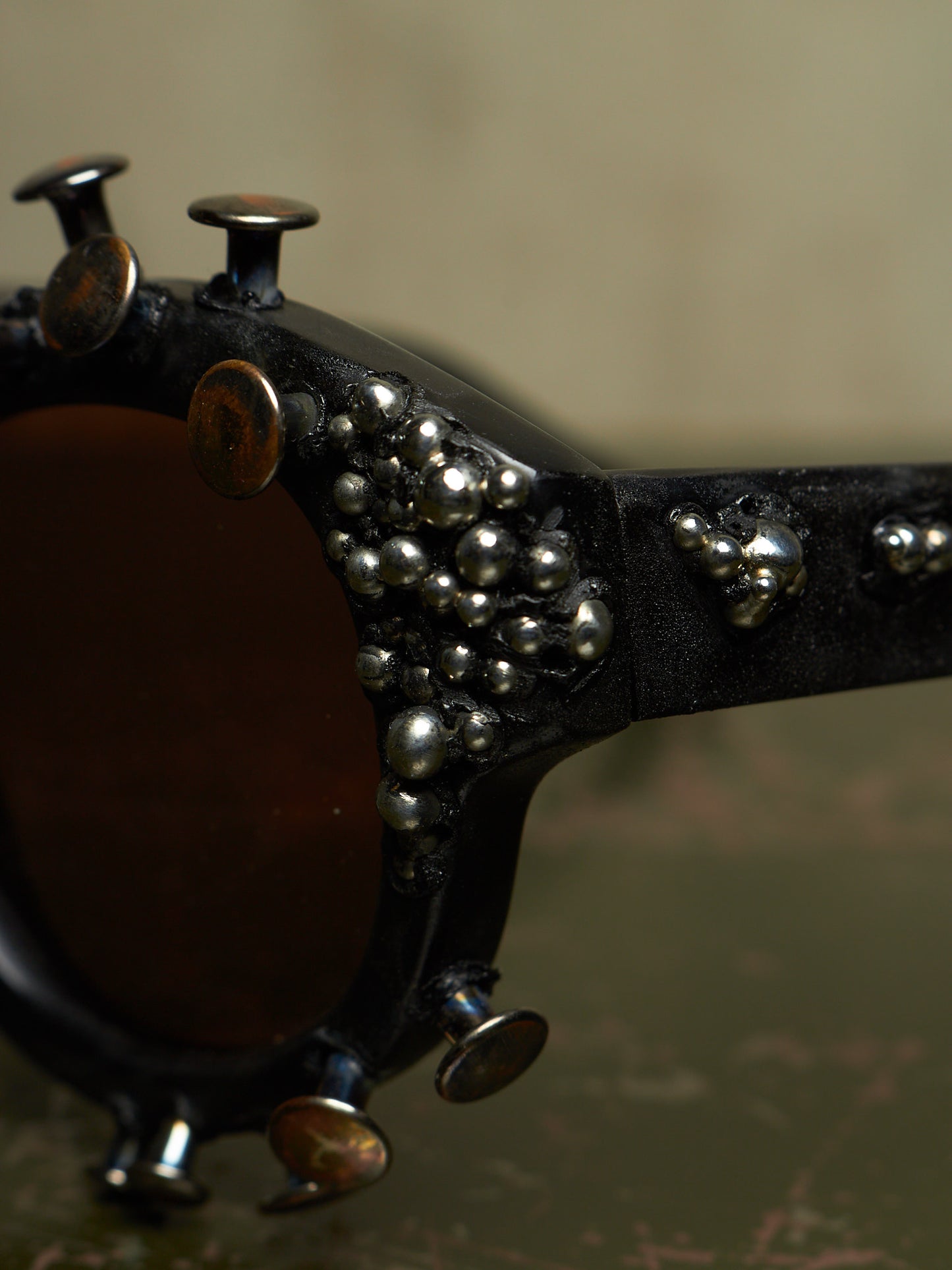 Close-up of a pair of sunglasses with a black frame heavily embellished with silver metallic studs, skull charms, and small beads, featuring dark brown lenses.