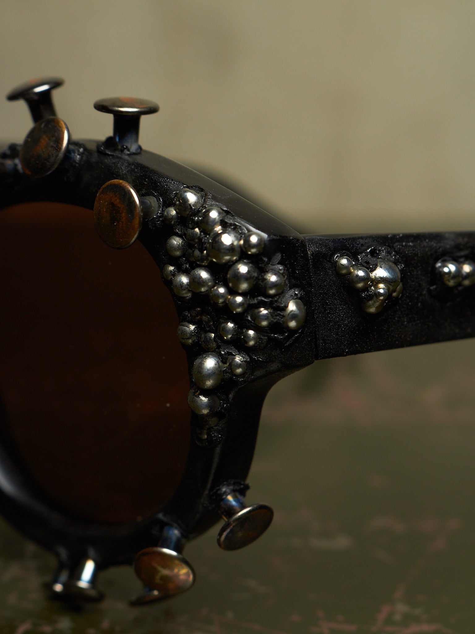Close-up of a pair of sunglasses with a black frame heavily embellished with silver metallic studs, skull charms, and small beads, featuring dark brown lenses.