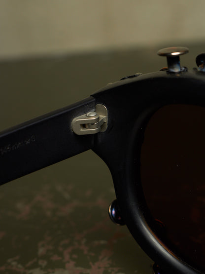 Close-up of a black sunglasses frame showing the temple arm, hinge hardware, and part of the dark lens.