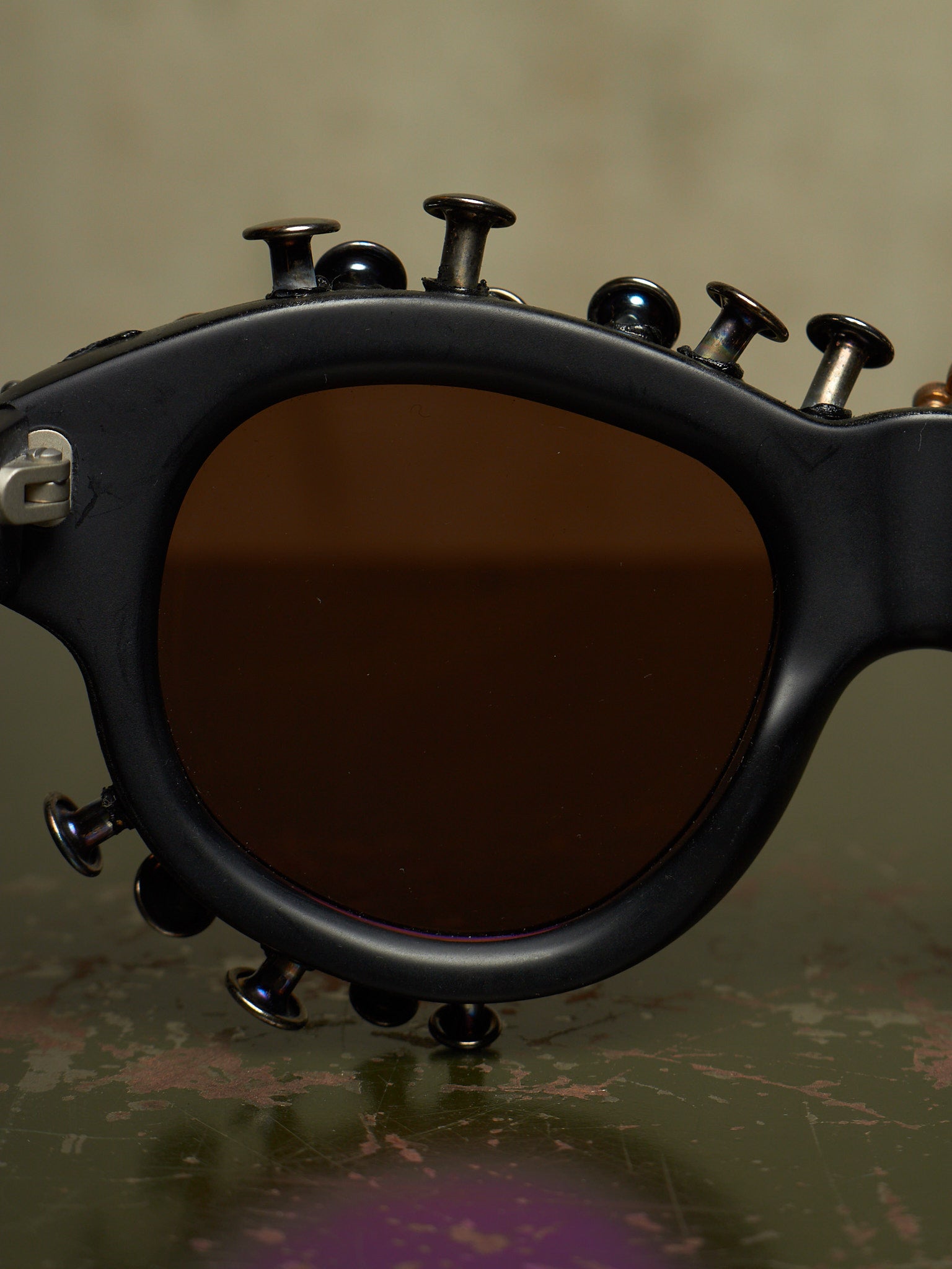 Close-up of a black novelty sunglasses frame with multiple metal screw-like embellishments around the edges and a dark brown tinted lens, resting on a textured dark surface.