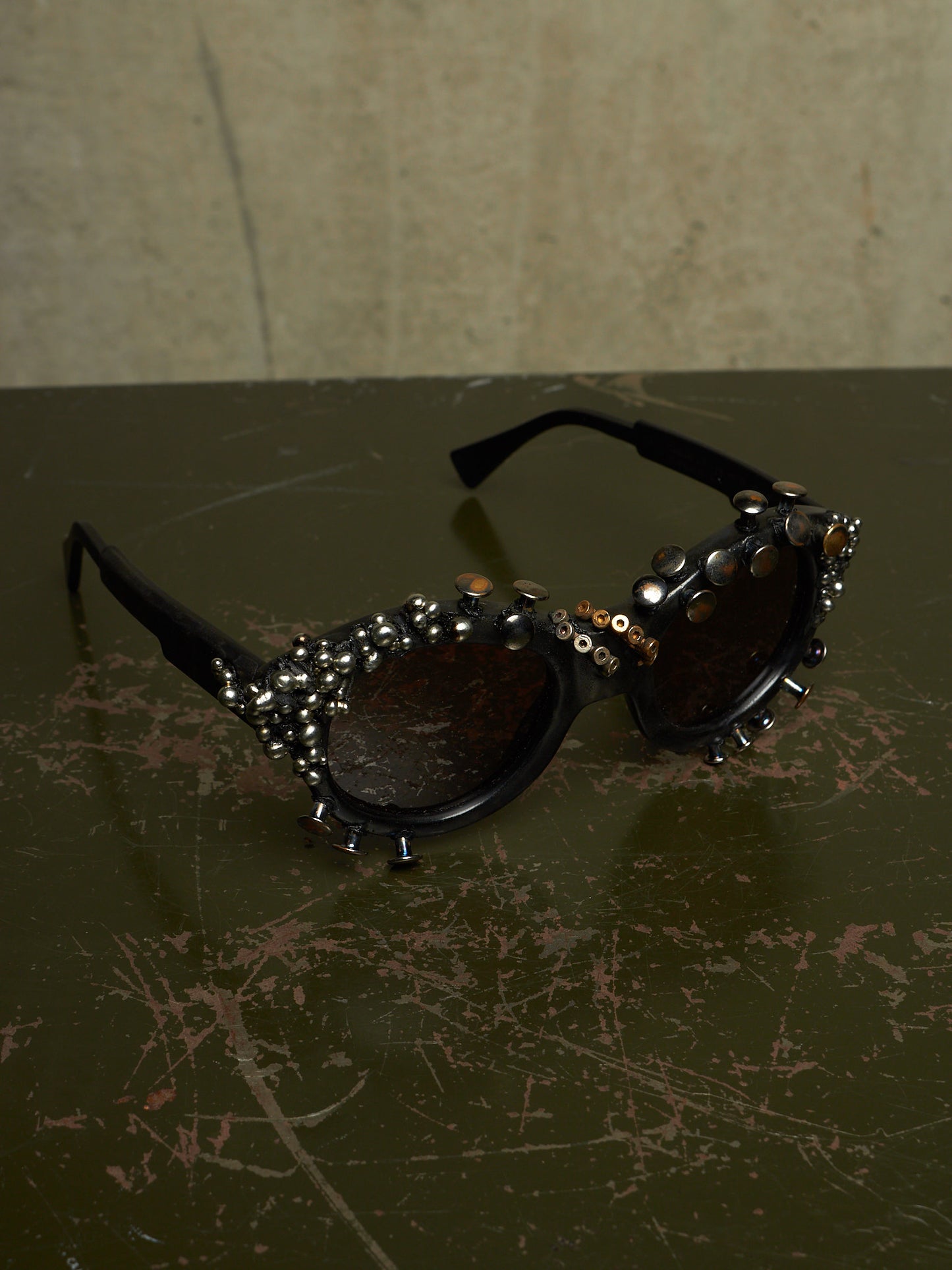 Black studded sunglasses with dark lenses and silver rhinestone embellishments, resting on a dark scratched tabletop against a textured beige wall.