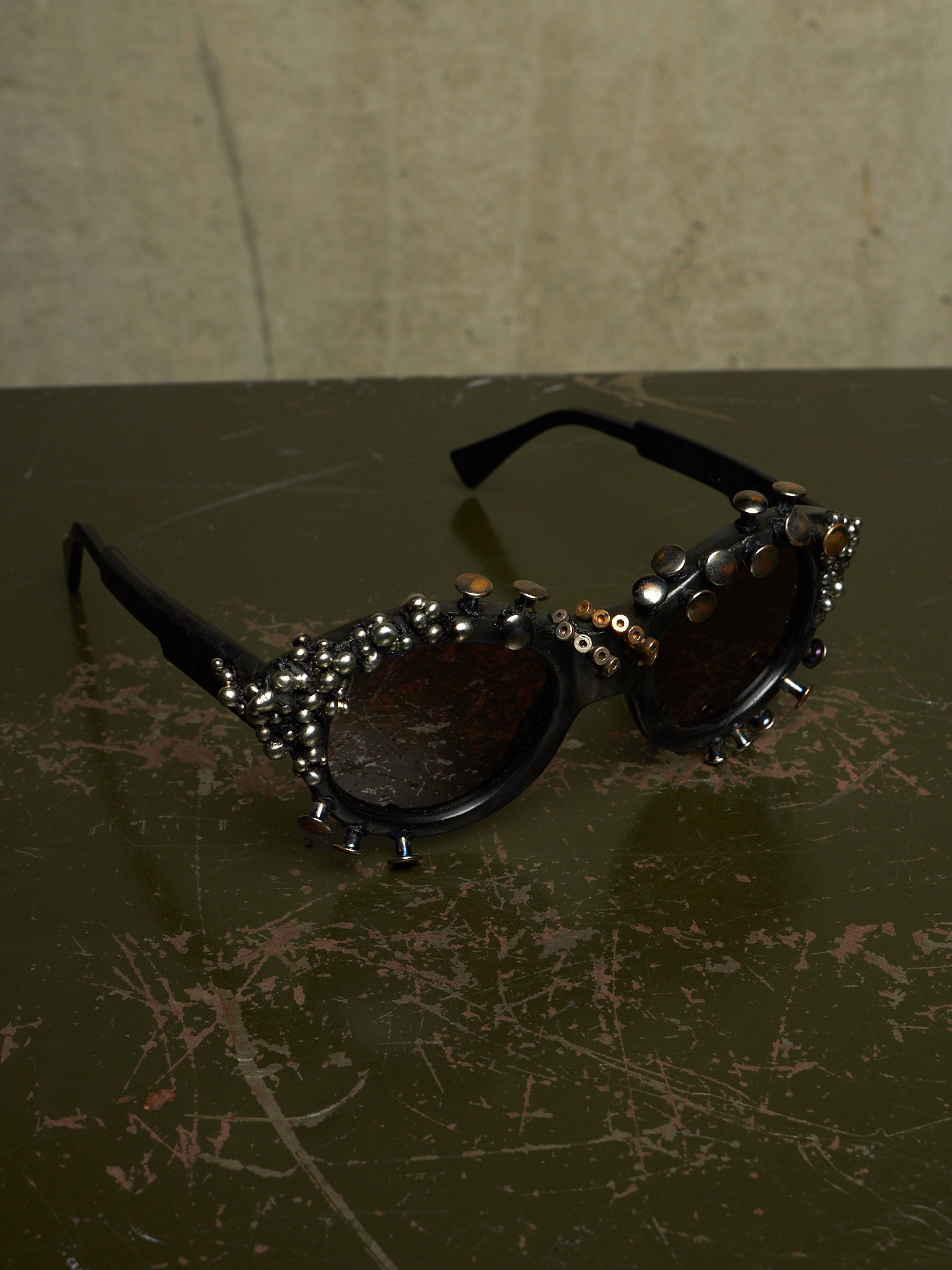 Black studded sunglasses with dark lenses and silver rhinestone embellishments, resting on a dark scratched tabletop against a textured beige wall.