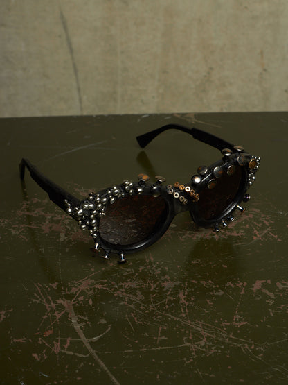 Black studded sunglasses with dark lenses and silver rhinestone embellishments, resting on a dark scratched tabletop against a textured beige wall.