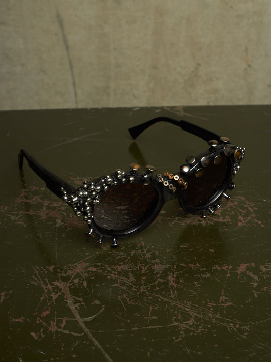 Black studded sunglasses with dark lenses and silver rhinestone embellishments, resting on a dark scratched tabletop against a textured beige wall.