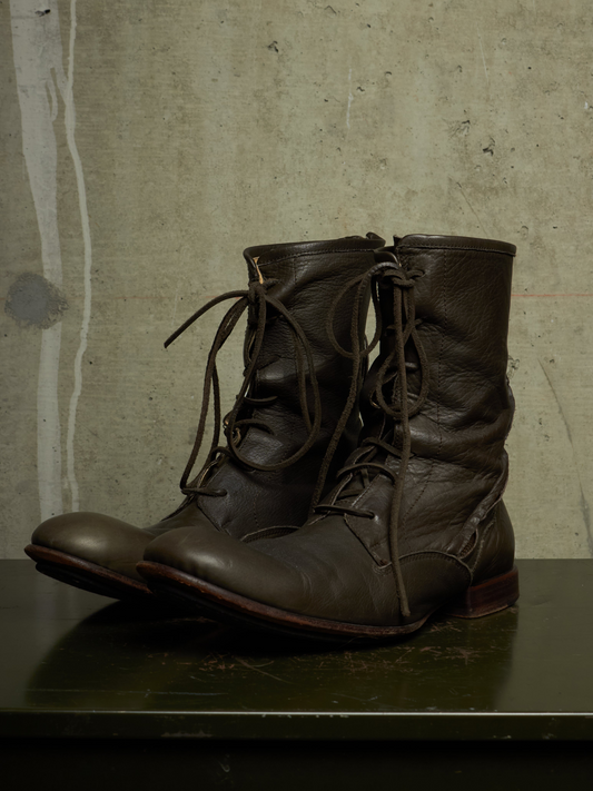 A pair of dark brown leather lace-up boots with a slightly slouchy, vintage silhouette, displayed on a dark surface against a textured concrete wall.