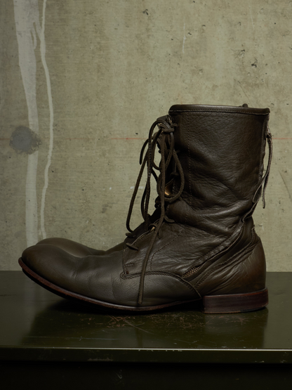 A dark brown leather lace-up ankle boot with a rugged, worn texture, resting on a dark green reflective surface against a textured concrete wall.
