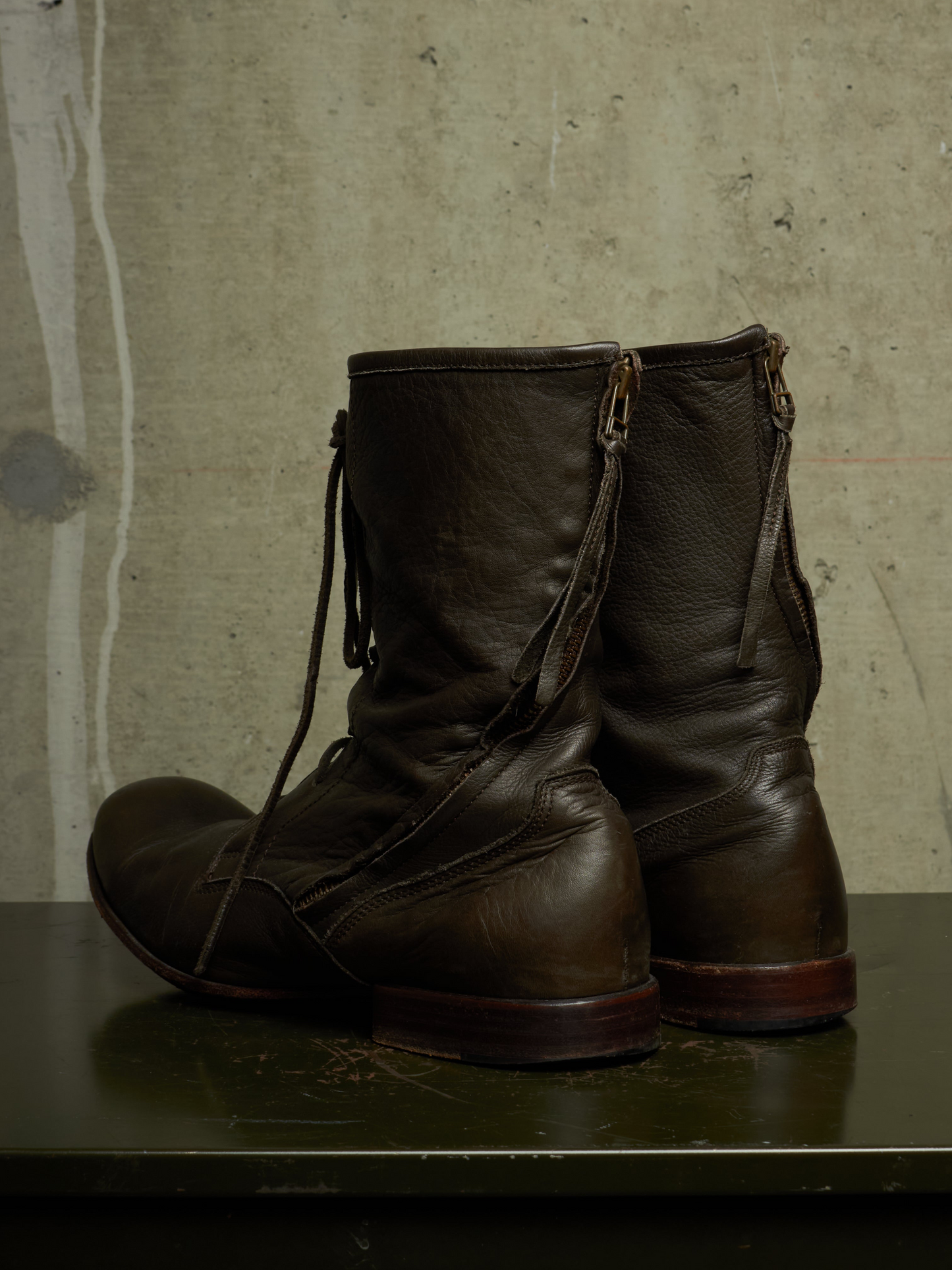 A pair of dark brown leather ankle boots with side zippers and lace-up fronts, displayed on a dark surface against a textured concrete wall.