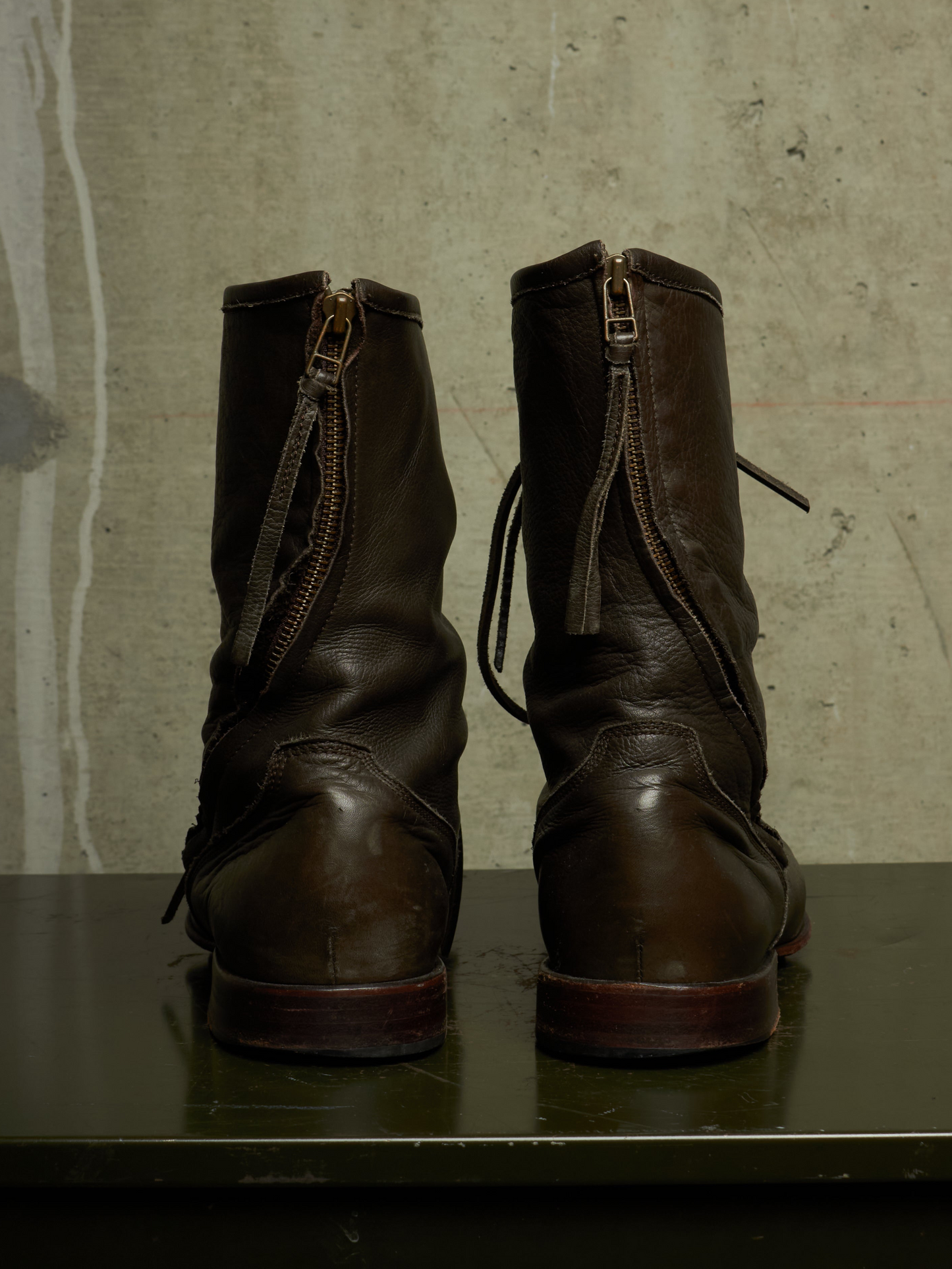 A pair of dark brown leather ankle boots with side zippers, shown from the back on a dark reflective surface against a textured concrete wall.