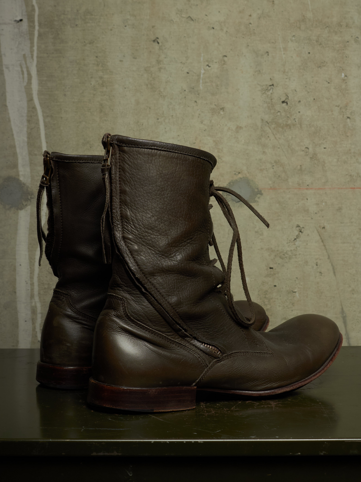 A pair of dark brown leather lace-up boots with side zippers, displayed on a dark green surface against a textured concrete wall.