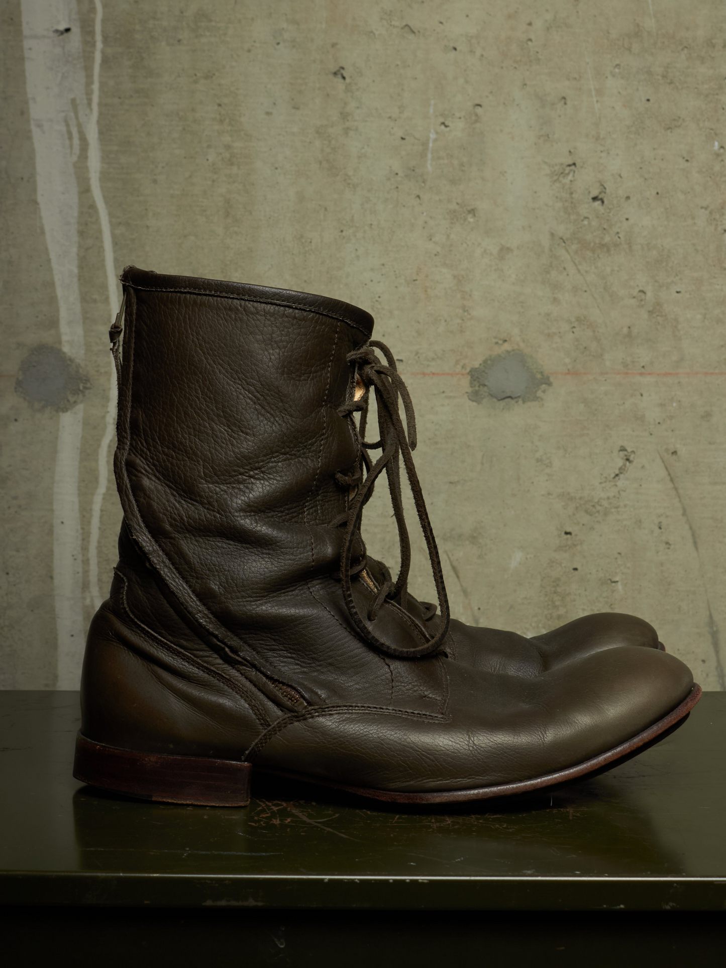 A pair of dark brown leather lace-up ankle boots resting on a dark green surface against a textured concrete wall.