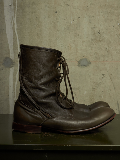 A pair of dark brown leather lace-up ankle boots resting on a dark green surface against a textured concrete wall.