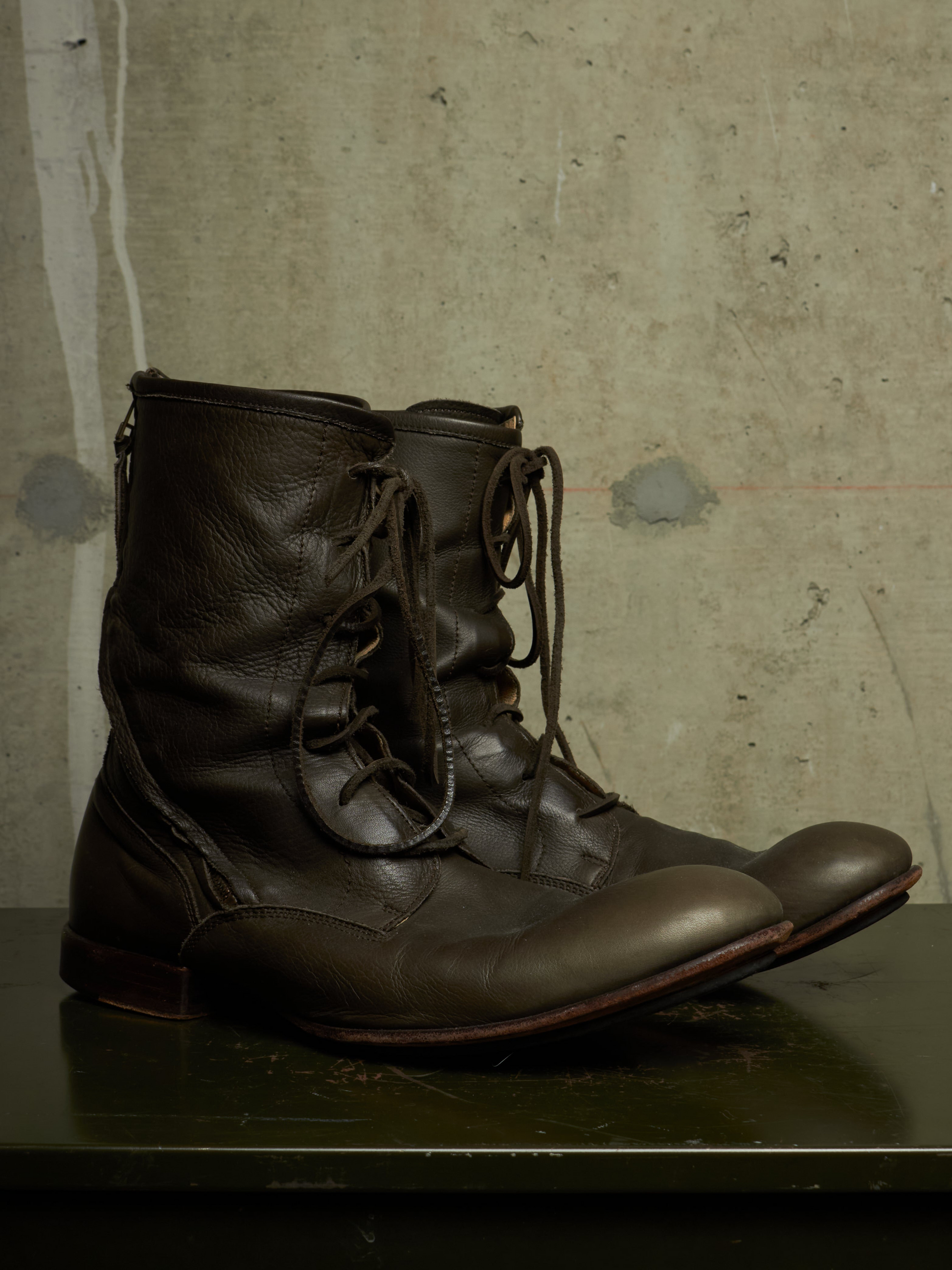 A pair of dark brown leather lace-up boots with a rugged, utilitarian design, resting on a dark surface against a textured concrete wall.