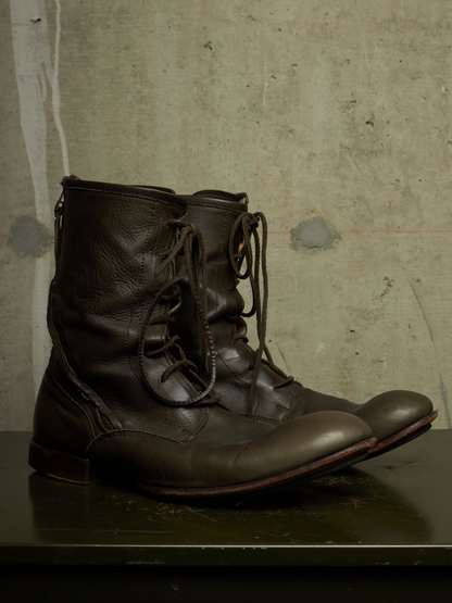 A pair of dark brown leather lace-up boots with a rugged, utilitarian design, resting on a dark surface against a textured concrete wall.