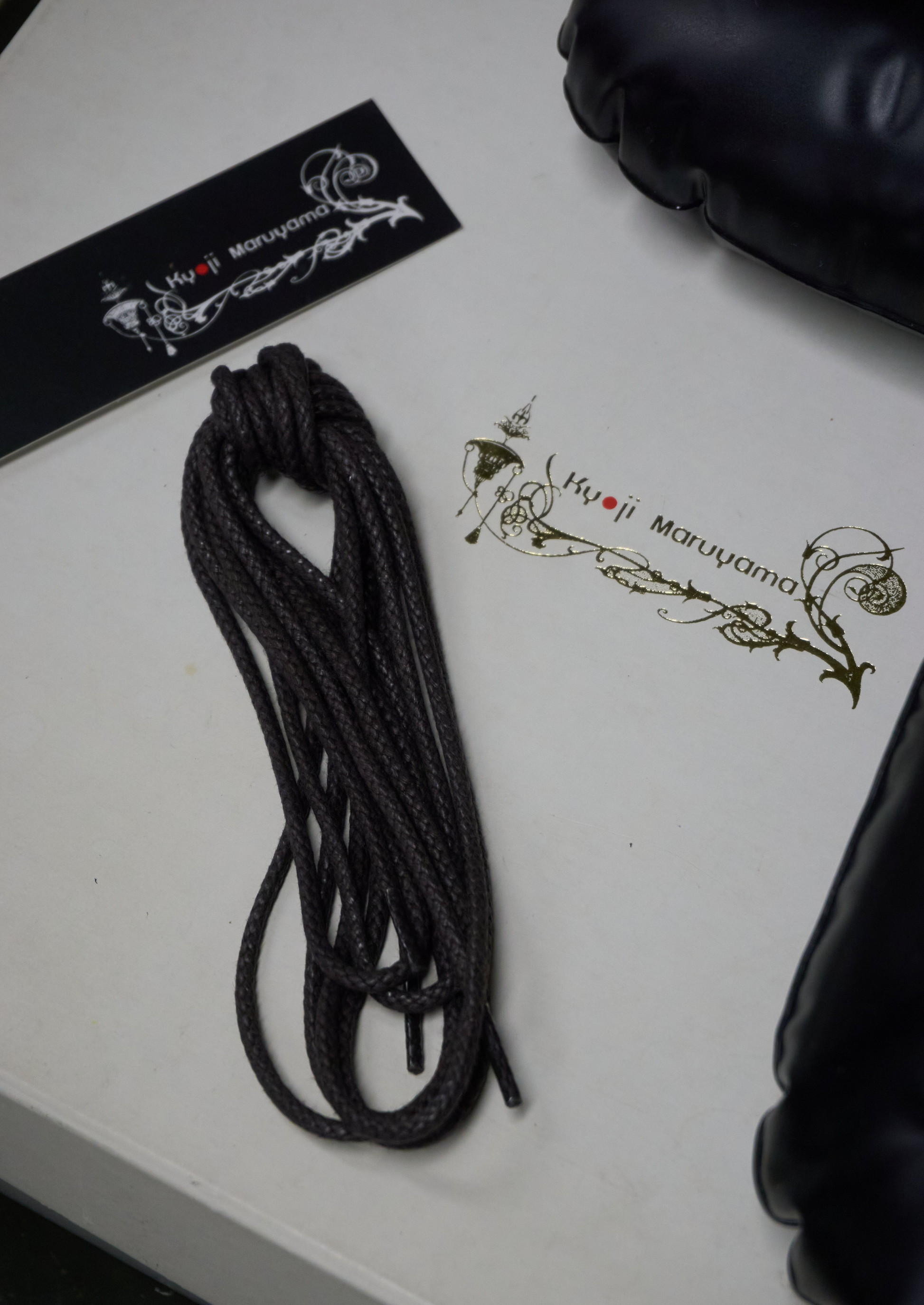 A coiled black rope or strap laid on a white surface next to branded packaging and a black padded item partially visible at the edges. The packaging shows the visible brand text 'Hijji Maruyama'.