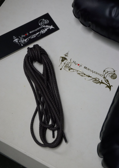 A coiled black rope or strap laid on a white surface next to branded packaging and a black padded item partially visible at the edges. The packaging shows the visible brand text 'Hijji Maruyama'.