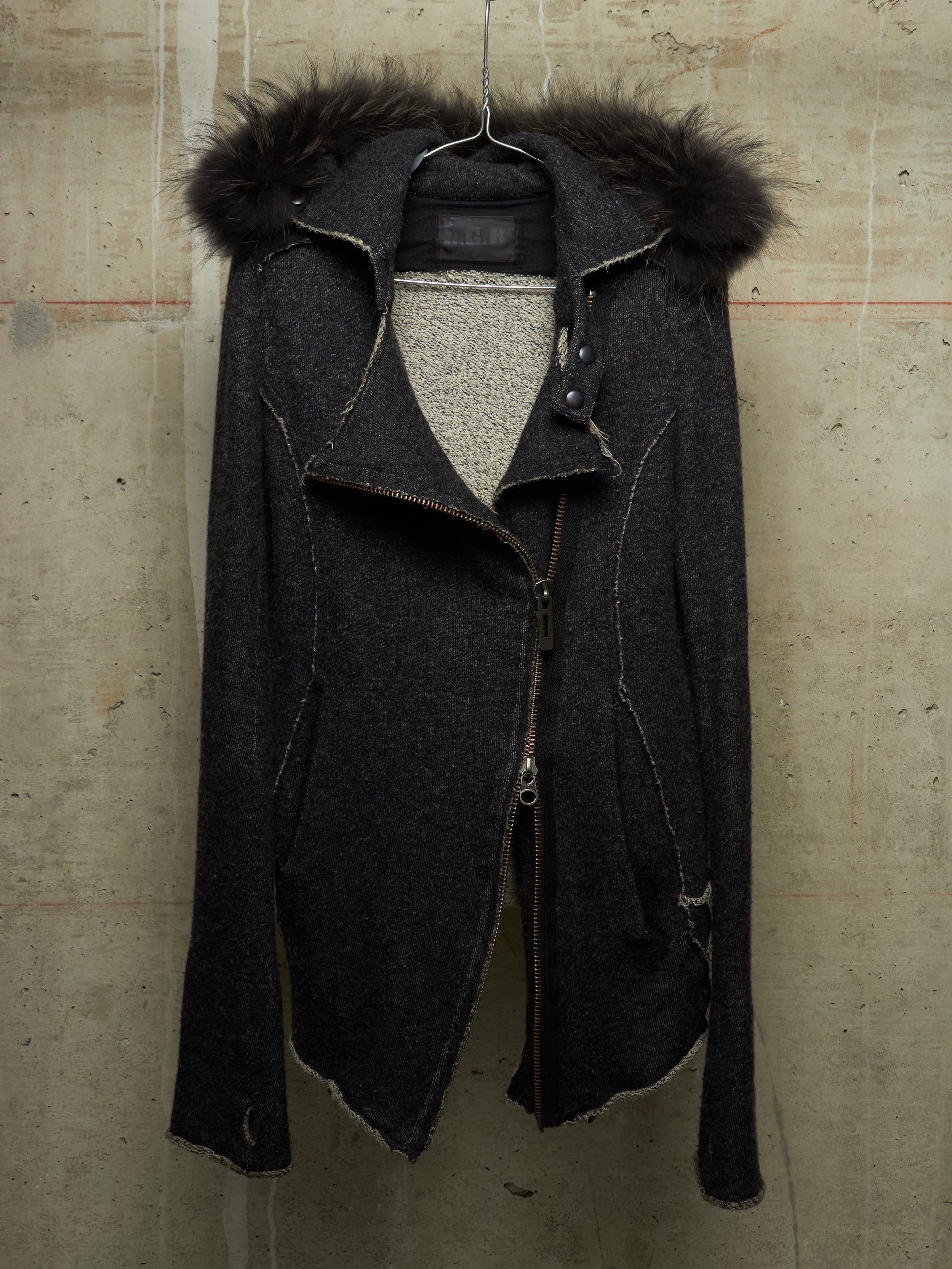 A dark charcoal gray asymmetrical zip-front jacket with a black fur-trimmed hood, hanging against a textured concrete wall.