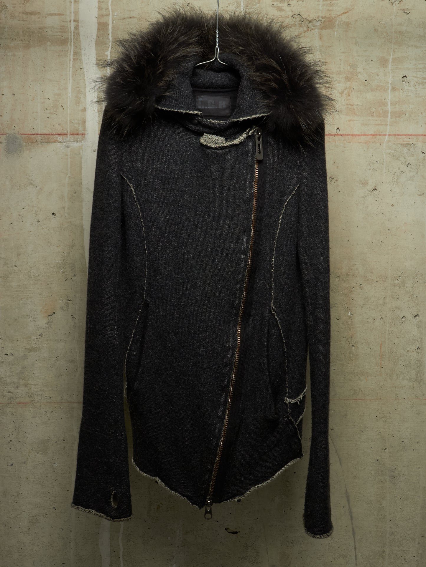 A dark charcoal gray asymmetrical zip jacket with a large dark brown fur-trimmed hood, hanging against a textured concrete wall.