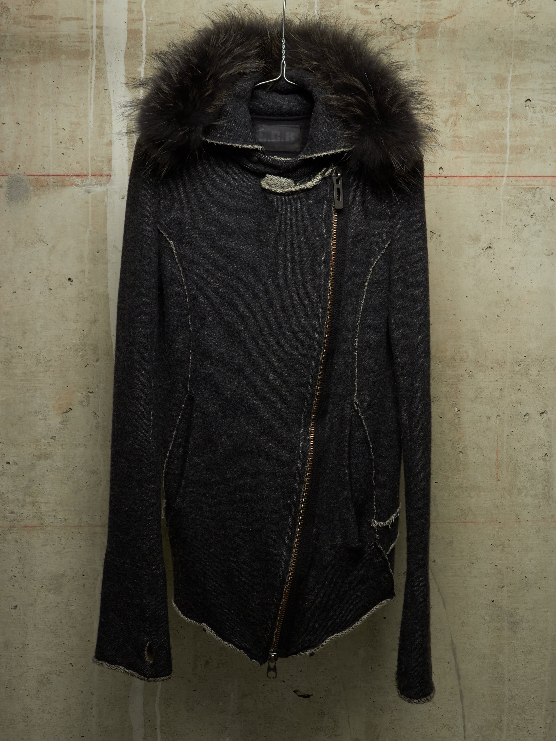 A dark charcoal gray asymmetrical zip jacket with a large dark brown fur-trimmed hood, hanging against a textured concrete wall.