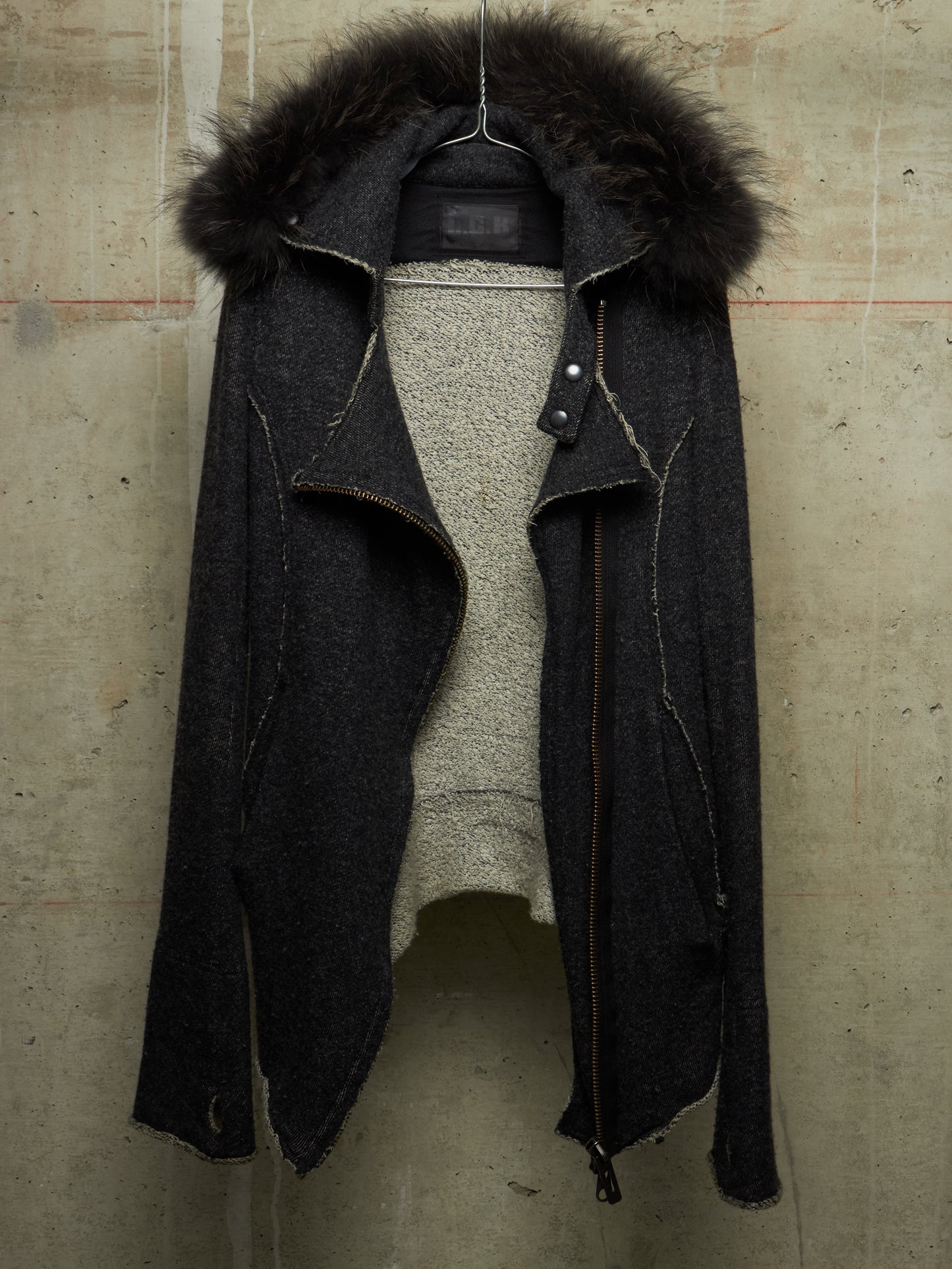A dark charcoal gray wool-blend jacket with a large black fur-trimmed hood, displayed open over a light gray knit sweater on a hanger against a textured concrete wall.