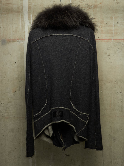 A dark charcoal gray knit cape-style cardigan with a thick black fur collar, photographed against a textured concrete wall.