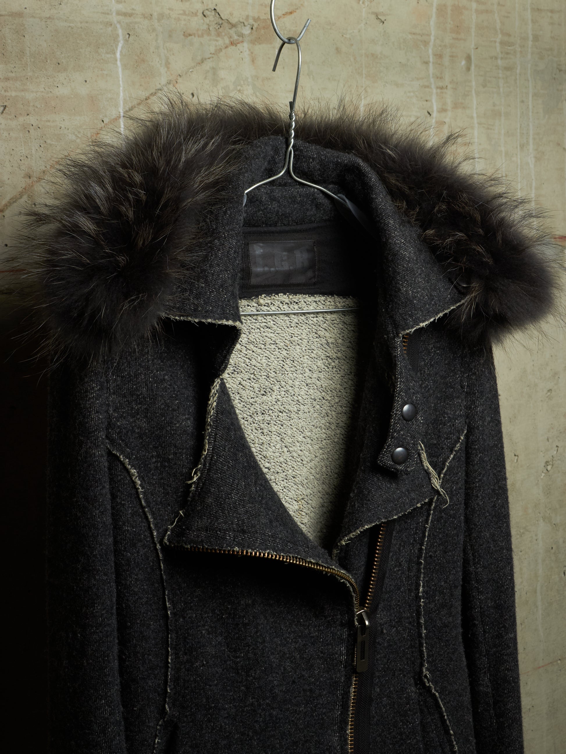 A dark charcoal gray wool coat with a large black fur-trimmed collar, hanging on a metal hanger against a textured beige wall.
