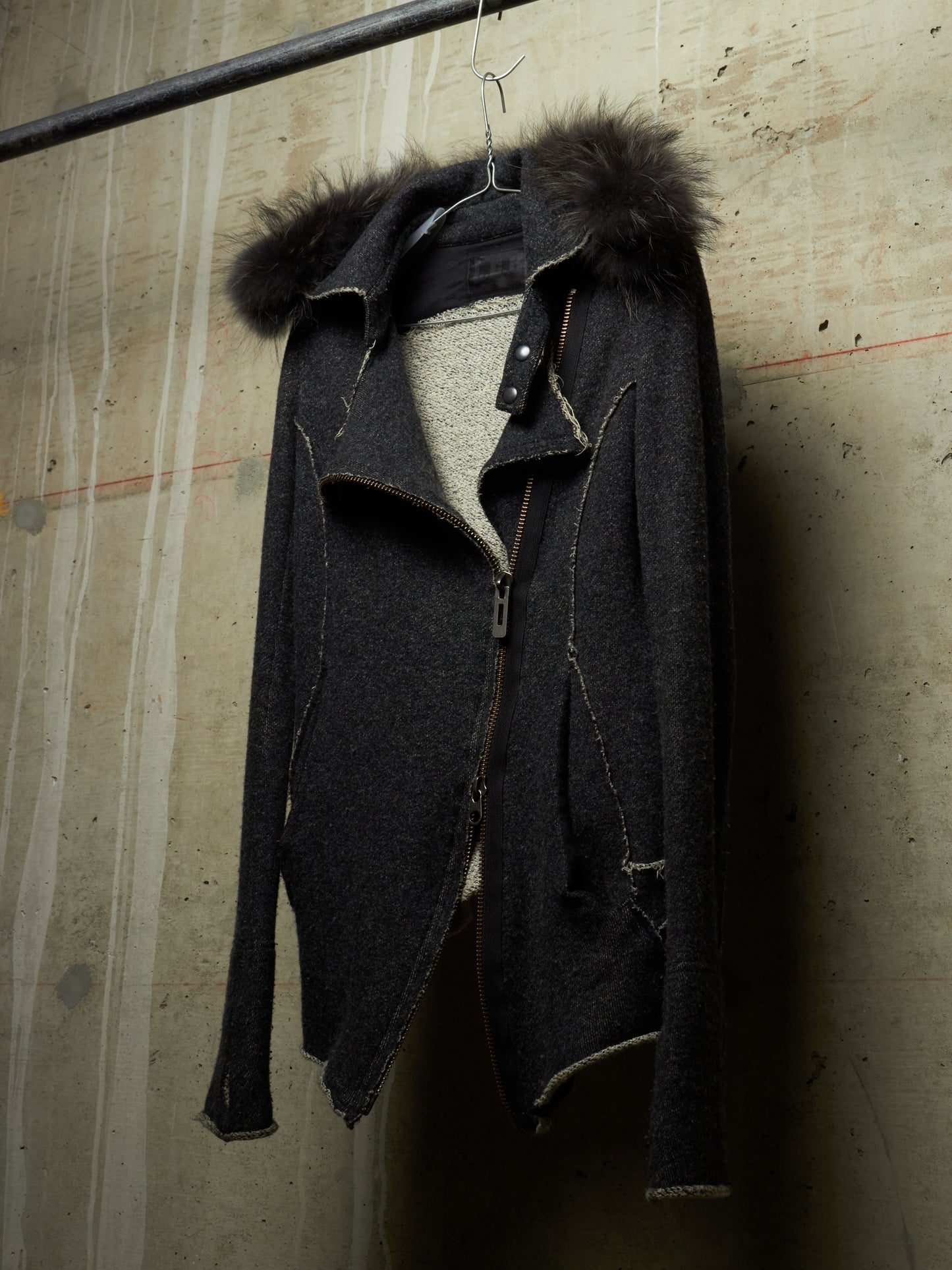 A dark charcoal zip-up jacket with a dark brown fur-trimmed hood, hanging on a metal hanger against a textured concrete wall.