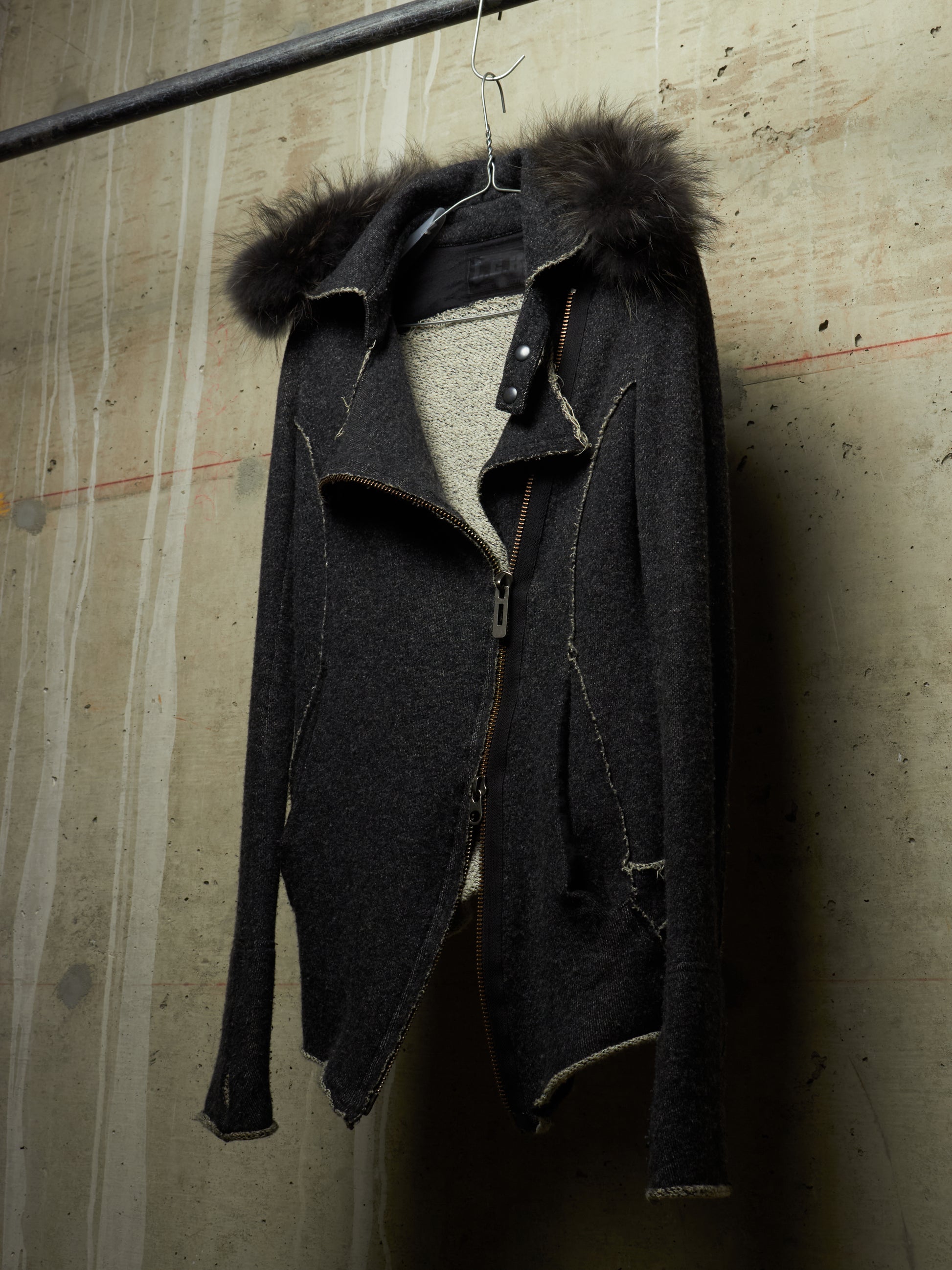 A dark charcoal zip-up jacket with a dark brown fur-trimmed hood, hanging on a metal hanger against a textured concrete wall.