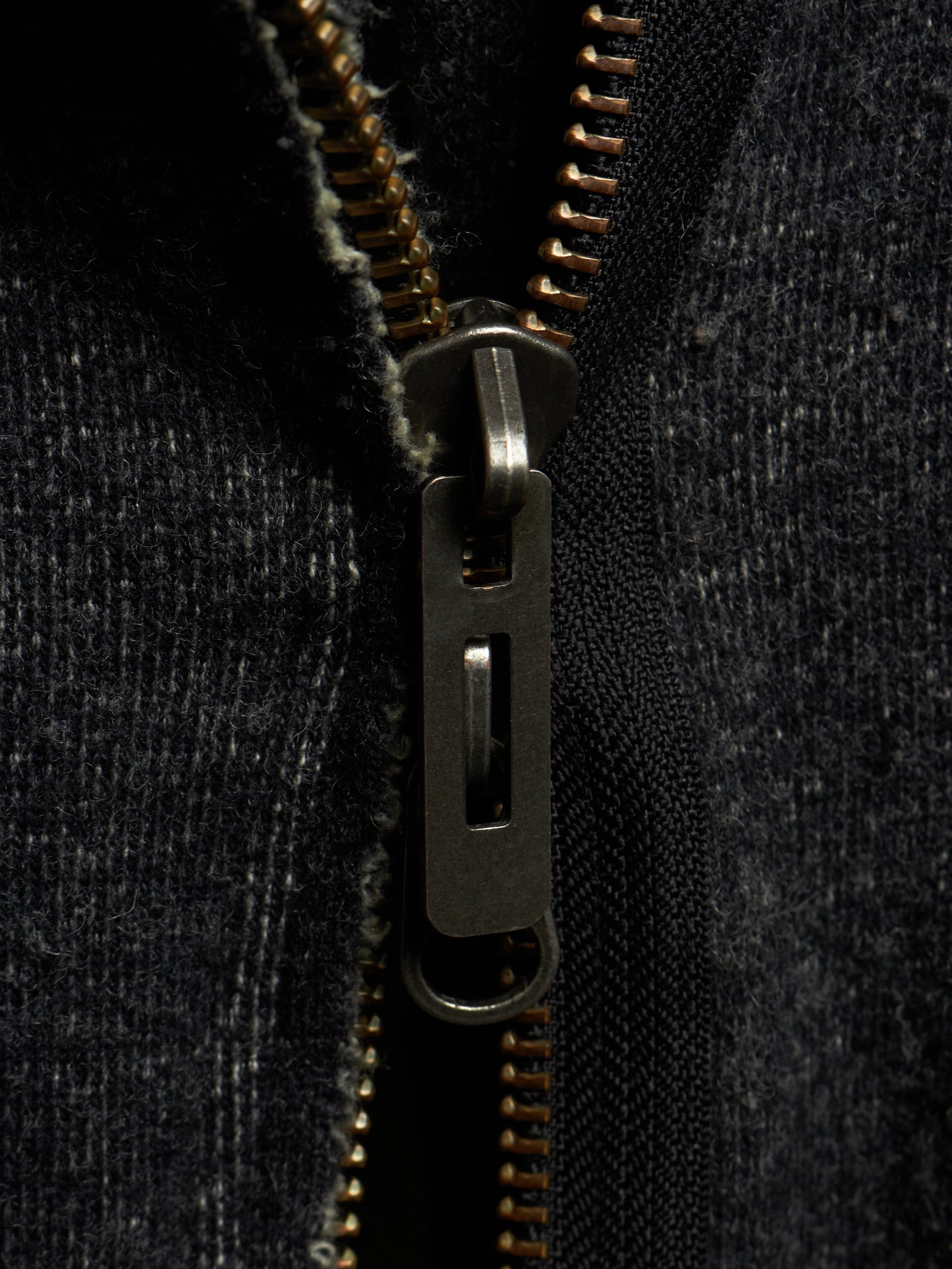 Close-up of a dark charcoal gray zippered garment with a dark metal zipper pull and gold-toned zipper teeth.