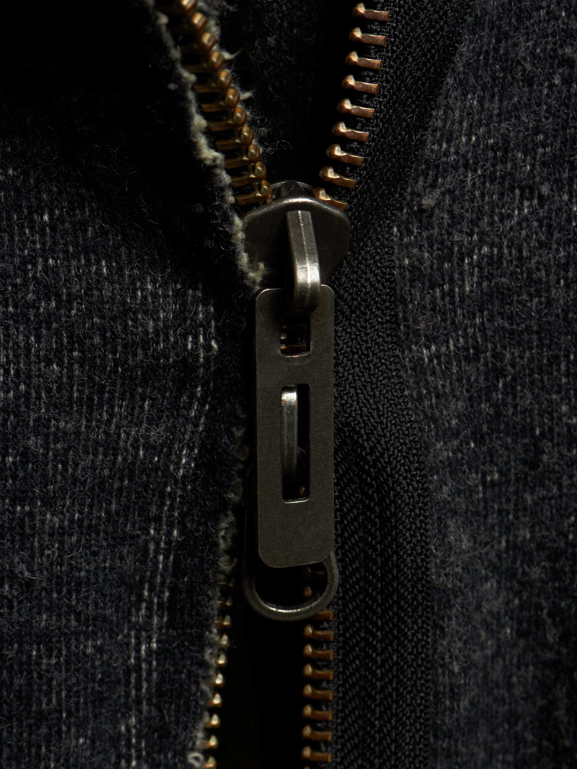 Close-up of a dark charcoal gray zippered garment with a dark metal zipper pull and gold-toned zipper teeth.