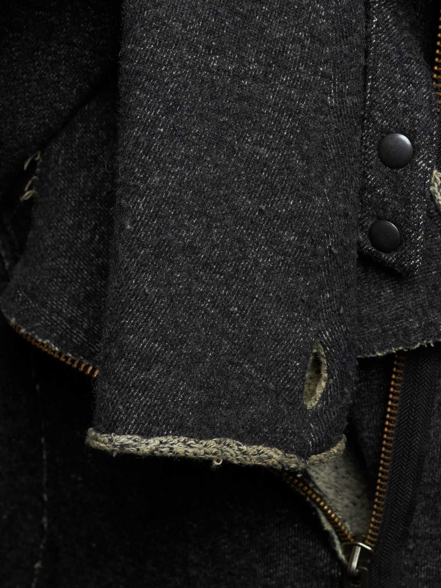 Close-up detail of a dark charcoal gray distressed denim jacket, showing the heavy textured fabric, raw frayed edges, metal snap buttons, and a brass zipper.