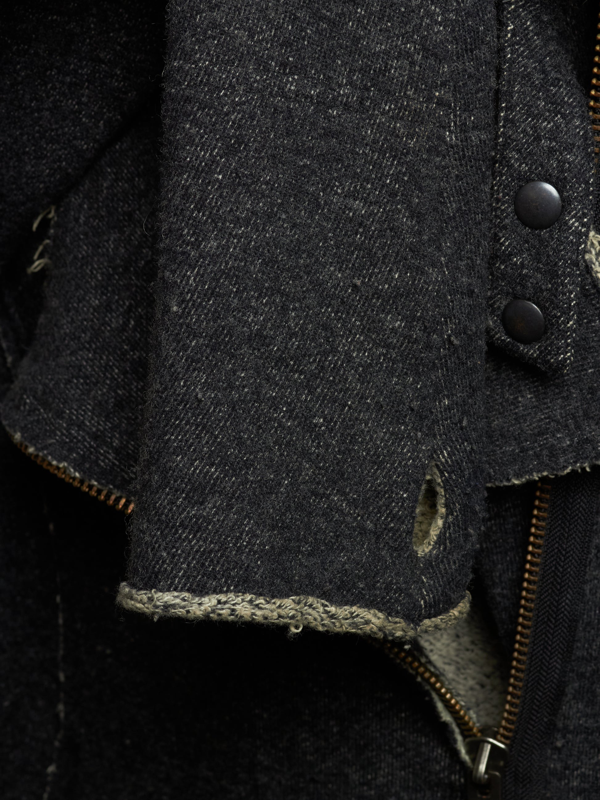 Close-up detail of a dark charcoal gray distressed denim jacket, showing the heavy textured fabric, raw frayed edges, metal snap buttons, and a brass zipper.