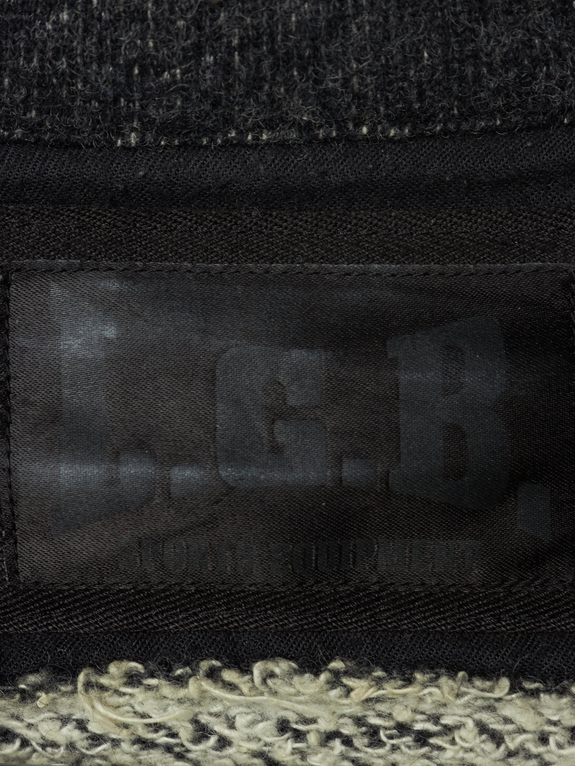 Close-up of a black woven fabric item with a dark translucent patch showing embossed letters and a textured cream and black fuzzy trim along the bottom edge.