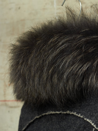 Close-up of a dark brown and black fluffy faux fur trim on a dark gray garment, hanging on a metal hanger against a light beige textured wall.