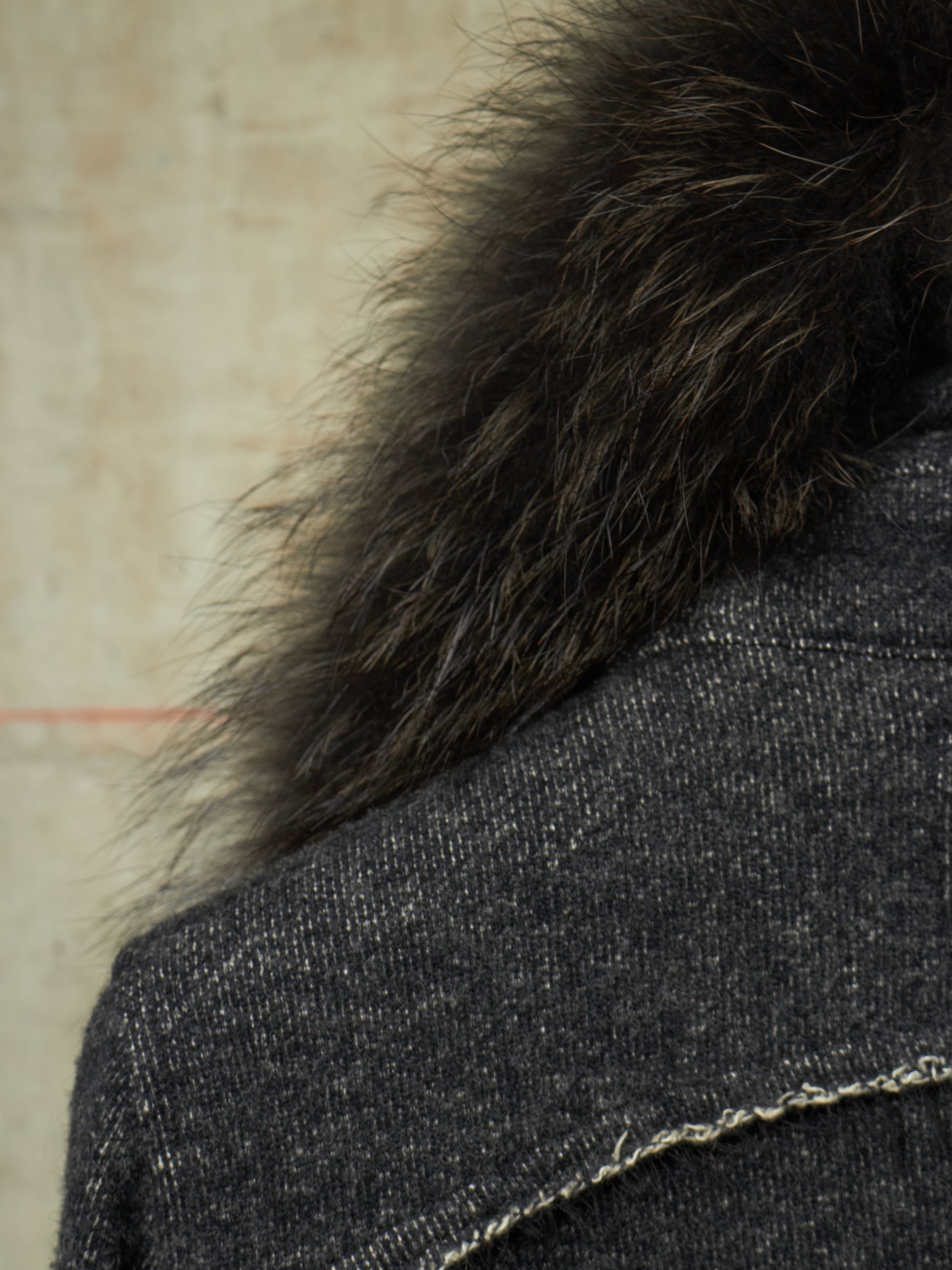 Close-up detail of a dark charcoal tweed jacket with a thick, fluffy black fur collar against a neutral beige background.