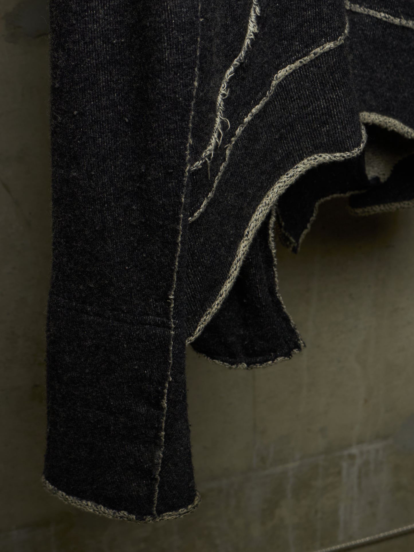 A close-up of a dark charcoal gray or black textured fabric garment, possibly denim or heavy cotton, featuring prominent light beige contrast stitching and raw, frayed edges, hanging against a neutral gray background.