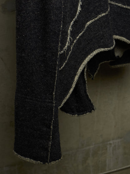 A close-up of a dark charcoal gray or black textured fabric garment, possibly denim or heavy cotton, featuring prominent light beige contrast stitching and raw, frayed edges, hanging against a neutral gray background.