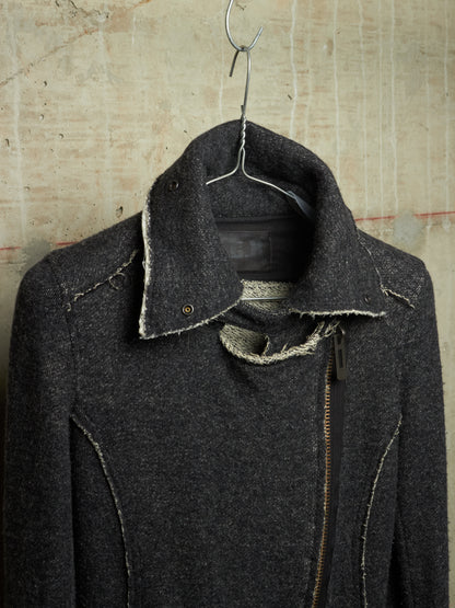 A dark charcoal gray textured jacket with raw-edge detailing and a front zipper, hanging on a metal hanger against a textured concrete wall.