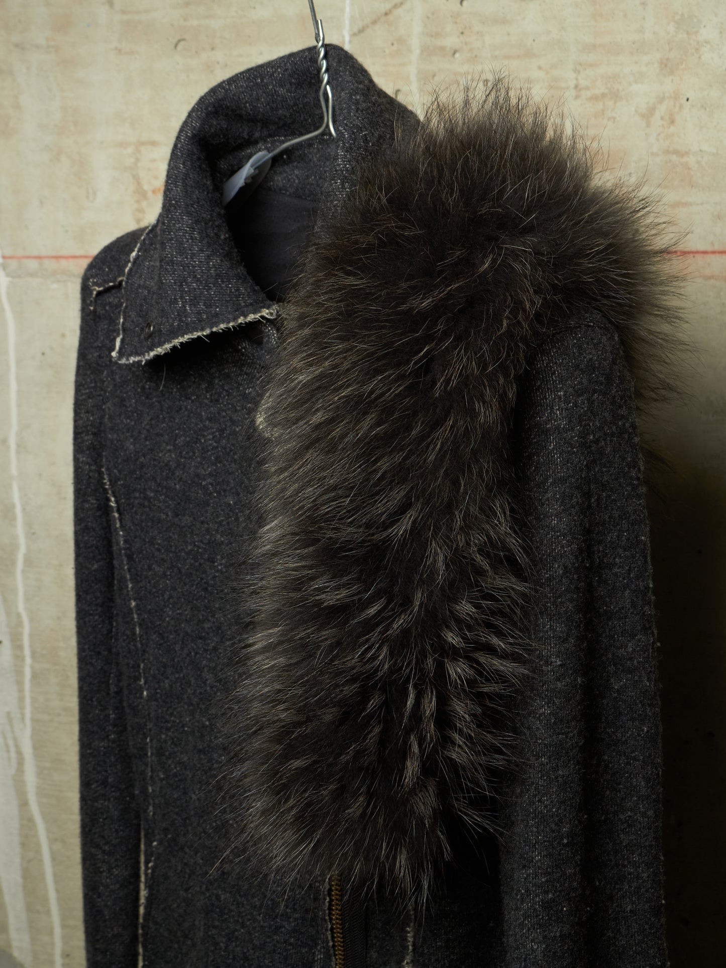 A dark charcoal gray wool coat with a large, fluffy black fur collar, hanging on a wire hanger against a textured beige wall.