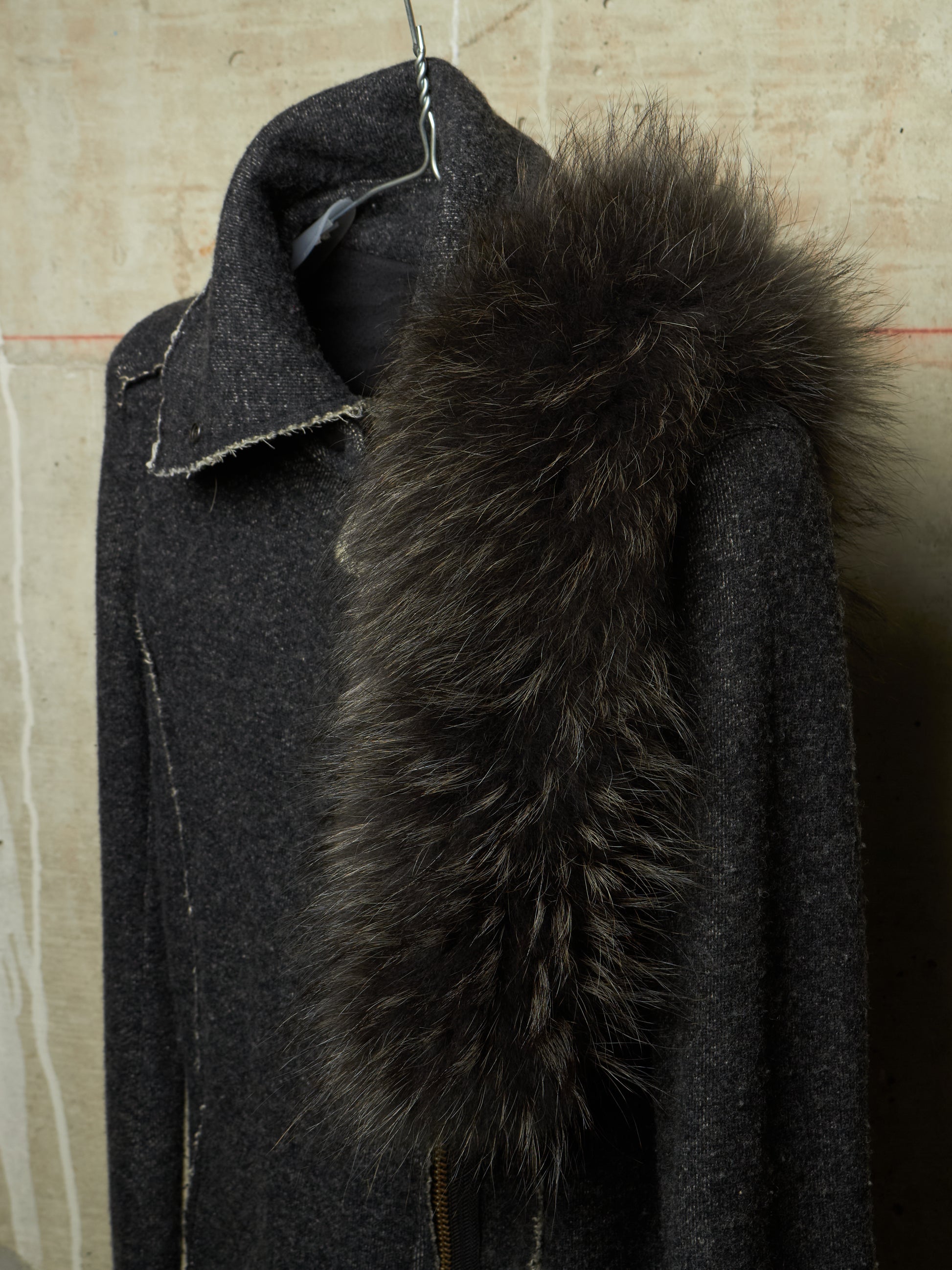 A dark charcoal gray wool coat with a large, fluffy black fur collar, hanging on a wire hanger against a textured beige wall.